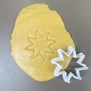 Può includere: Un tagliabiscotti bianco a forma di stella accanto a un pezzo di pasta gialla con un'impronta a stella. L'impasto è su una superficie grigia. Il tagliabiscotti ha un bordo frastagliato.