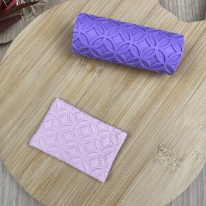 Peut inclure: Un rouleau à pâtisserie cylindrique violet avec un motif géométrique en relief de cercles entrelacés et une plaque rectangulaire violet clair assortie présentant le même motif circulaire gaufré pour créer des impressions texturées.