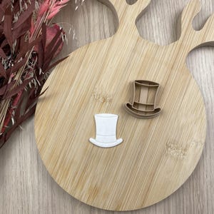 May include: A white top hat shaped cookie cutter and a matching white top hat shaped cutout piece. The cookie cutter is tan and features a distinct brim and crown structure, while the cutout piece is smooth and solid white in color.