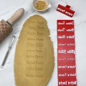Op de afbeelding: Een houten deegroller met bloemmotief, een metalen garde, een kommetje met gouden strooisels en een lap deeg met de tekst "Votre Texte" erin gedrukt. Naast het deeg liggen diverse rode rechthoekige stempels met de tekst "Votre Texte".