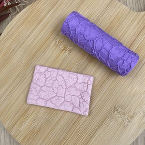 Peut inclure: Un rouleau cylindrique violet avec un motif en relief de fissures irrégulières et une plaque rectangulaire en pâte lavande claire présentant le même motif texturé, utilisés pour imprimer des textures sur des matériaux souples.
