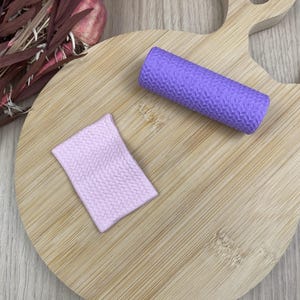 Op de afbeelding: Een cilindrische paarse roller met een gestructureerd visgraatpatroon en een rechthoekig lichtroze vel met hetzelfde reliëf visgraatontwerp, beide rustend op een ronde houten plank met handvat, naast gedroogde florale elementen.