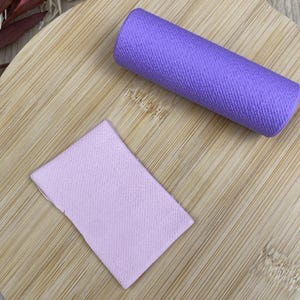 May include: A bright purple fabric roll and a rectangular piece of light pink fabric. Both items feature a visible woven texture and are positioned on a light-colored wooden surface with a rounded shape and a handle.