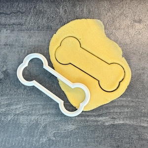 Peut inclure: Un emporte-pièce en plastique blanc en forme d'os pour chien à côté d'un morceau de pâte jaune avec une forme d'os découpée. Le découpoir est sur une surface grise.