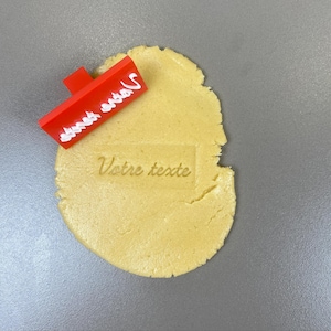 May include: A red cookie stamp with white text reading "Votre texte" is pressed into a piece of dough. The stamp is rectangular with a handle. The dough is light yellow and has the same text imprinted on it.