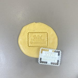 May include: A light yellow dough with a rectangular imprint that reads "Bébé EN COURS" and a heart symbol. A white plastic cookie cutter with the same text and design is next to the dough. The dough is on a gray surface.