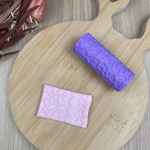 May include: A purple cylindrical rolling pin with a raised cow print pattern and a matching light purple rectangular clay slab featuring the same embossed cow print texture, designed for creating consistent patterns on soft crafting materials.