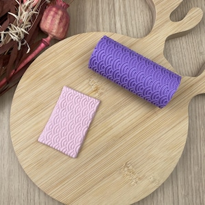 Peut inclure: Un rouleau à pâtisserie violet et un petit outil rectangulaire rose, tous deux avec un motif de vagues répétées, reposent sur une planche à découper en bois. Le rouleau est cylindrique et l'outil est plat. Des fleurs séchées et une gousse décorative sont en arrière-plan.