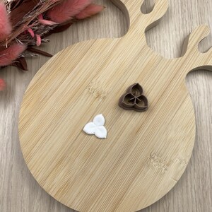 May include: A brown plastic three-petaled flower cookie cutter and a matching white fondant cutout piece. The cutter features a central Y-shaped divider to create petal impressions, while the white cutout shows the finished three-lobed shape.