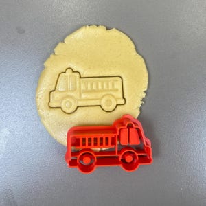 Peut inclure: Un emporte-pièce en plastique rouge en forme de camion de pompiers à côté d'un morceau de pâte avec une empreinte de camion de pompiers. Le découpoir présente des détails, notamment une échelle et des roues. La pâte est de couleur beige clair, et le fond est gris uni.