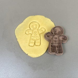 Puede incluir: Un cortador de galletas con forma de hombre de jengibre junto a una forma recortada en masa. El cortador es marrón y tiene forma de hombre de jengibre con los brazos extendidos. La masa es de color amarillo claro.