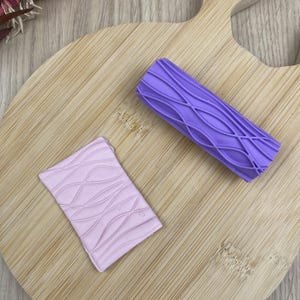 Puede incluir: Una herramienta de rodillo cilíndrico de color morado con un patrón de celosía en relieve y una placa de arcilla rectangular de color rosa claro grabada con el mismo diseño de ondas entrelazadas, mostrando la textura creada por el rodillo.