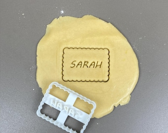 May include: A light-colored dough with the word "SARAH" imprinted in a rectangular shape. A white plastic cookie cutter with the word "SARAH" in reverse is next to the dough. The cutter has a scalloped edge.