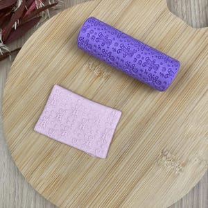 Peut inclure: Un rouleau à pâtisserie cylindrique violet avec un motif en relief de bonshommes en pain d'épice et de petites fleurs, accompagné d'un morceau rectangulaire d'argile violet clair présentant le même motif de bonshommes et de fleurs.
