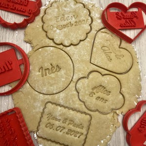 Op de afbeelding: Een vel rauw koekjesdeeg met verschillende rode plastic koekjesvormpjes erin gedrukt. Het deeg bevat reliëftekst met namen zoals Eden, Inès, Alice en Yann & Paola, samen met datums en de tekst 6 ans, wat zorgt voor gepersonaliseerde vormen.