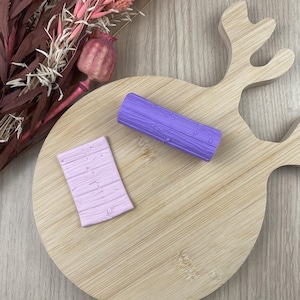 May include: Two small, textured, wood-grain-patterned clay pieces in lavender and light pink rest on a wooden cutting board. Dried pink and burgundy flowers and seed pods are in the background. The cutting board has a deer head shape.