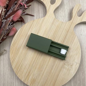 Peut inclure: Un étui coulissant rectangulaire vert olive avec une piste intérieure nervurée et un petit composant cylindrique blanc positionné à l'intérieur de la piste. L'étui présente une finition mate lisse et un mécanisme coulissant fonctionnel.
