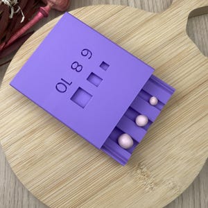 May include: A lilac-coloured sliding box with the numbers 6, 8, and 10 printed on the side. The box contains three small, pink spheres. The box is on a wooden chopping board.