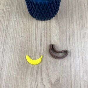 Peut inclure: Un banane jaune et un emporte-pièce en forme de banane marron sont posés sur une surface en bois. Un pot géométrique bleu foncé avec une petite plante est en arrière-plan. La banane est jaune vif.
