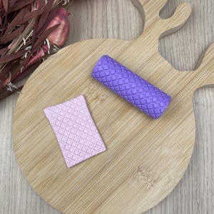 Peut inclure: Un rouleau cylindrique violet et une plaque rectangulaire violet clair, tous deux dotés d'un motif en relief en forme de grille. Ces objets sont placés sur une planche en bois clair en forme de tête de cerf, avec des fleurs séchées en haut à gauche.
