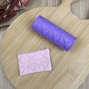 May include: A cylindrical purple rolling pin with a raised geometric lattice pattern and a matching rectangular light purple clay slab featuring the same embossed design, showcasing a repeating network of lines and small circular nodes.