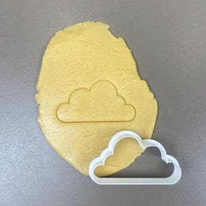 Può includere: Un tagliabiscotti bianco a forma di nuvola accanto a un pezzo di pasta cruda giallo chiaro. L'impasto ha una forma di nuvola premuta. Il tagliabiscotti è largo circa 10 cm.