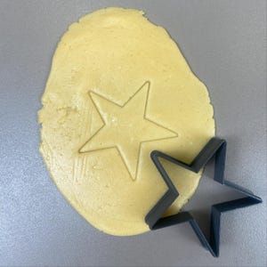 Peut inclure: Un emporte-pièce en forme d'étoile à côté d'un morceau de pâte à biscuits avec une empreinte d'étoile. La pâte est jaune clair et l'emporte-pièce est gris foncé. Vue de dessus.