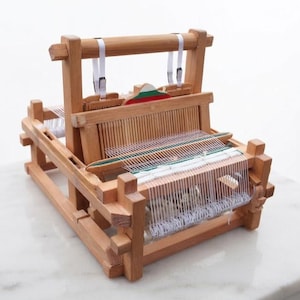 Pode incluir: Um tear de mesa de madeira com acabamento natural em castanho claro e uma urdidura de fio branco. O dispositivo inclui uma moldura de liço central com uma pequena peça decorativa que exibe faixas horizontais nas cores branca, verde e vermelha.