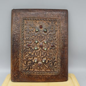 Puede incluir: Un diario rectangular de cuero marrón con un diseño floral en relieve. El panel central presenta un intrincado patrón con adornos rojos y verdes. El diario se exhibe sobre una superficie de madera clara.