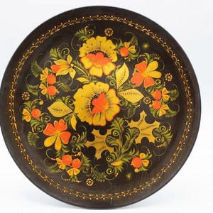 Könnte beinhalten: Ein runder, schwarzer Zierteller mit einem handgemalten Blumenmuster in Gelb-, Orange- und Grüntönen. In der Mitte befinden sich große Blüten, umgeben von kleineren Blumen und Blättern, sowie ein feiner, gepunkteter Rand entlang der Kante.
