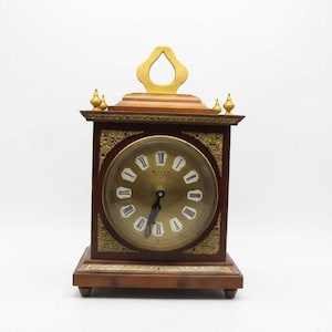 May include: A wooden mantel clock with a dark brown finish, featuring a circular brass-toned face with Roman numerals and the text "WEHRLE". The clock includes ornate gold-colored metal corner accents, a top handle, and four small feet.