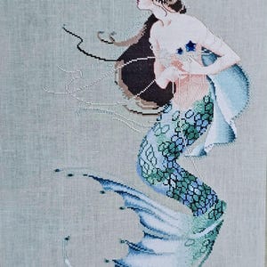 May include: A cross-stitch embroidery of a mermaid with long flowing hair, a blue and green tail, and a pearl necklace. The mermaid is holding a seashell in her hands.