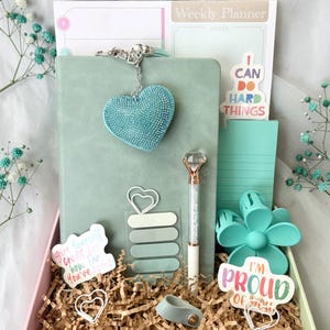 May include: A gift box filled with stationery items in shades of teal and pink. Includes a journal, pen, sticky notes, stickers with text, and decorative paperclips. A teal heart-shaped charm hangs from the journal. The words "I CAN DO HARD THINGS" are visible.