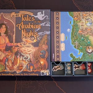 May include: Open board game box with the title "Tales of the Arabian Nights" and a 40th Anniversary logo. The box art features a woman in pink clothing. The game includes a map, game pieces, and a storage tray.