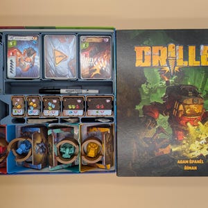 Peut inclure: Un jeu de société intitulé Drillers avec un couvercle illustré d'une foreuse rouge et une boîte ouverte contenant des composants organisés, dont des paquets de cartes, des jetons de ressources, des figurines et un marqueur noir.