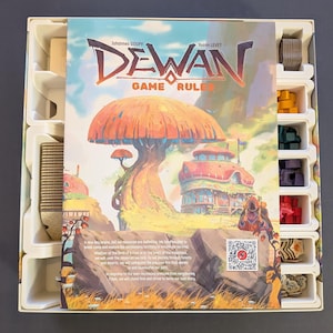 May include: A board game box with the title "DEWAN" in red and orange lettering. The game rules cover features a fantasy landscape with a large mushroom and colorful buildings. The box contains game pieces in various colors.