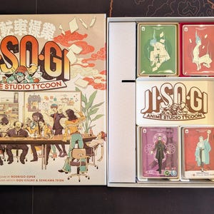 May include: The board game Ji-So-Gi Anime Studio Tycoon features a box lid with an illustration of an office scene and a game box interior containing organised card decks, a white tray with the game title, and three grey rectangular dividers.
