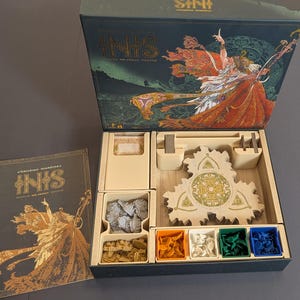 May include: Open board game box with the title "INIS" and artwork of a figure in orange and red robes. Inside are game pieces, including miniature figures and colored tokens. A separate book with the same artwork is visible.