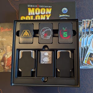 May include: A board game titled "Moon Colony" by Donald X. Vaccarino. The game box is black with gold and white text. Inside the box are cards with carbon fiber textures and various graphics. An instruction manual and game cards are also visible.