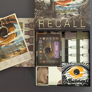 May include: Open board game box with the title "RECALL" in gold lettering. The box contains game components, including cards, tokens, and a game board with a spiral design. The box art features a landscape with a large eye.