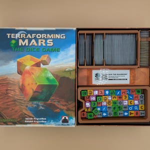 May include: Terraforming Mars The Dice Game board game box with lid open. The box cover features a Martian landscape and dice. Inside, a brown plastic insert holds stacks of cards, a white instructional sticker, and numerous colorful dice.