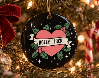 Op de afbeelding: Een ronde zwarte kerstornament van keramiek met een roze hartafbeelding in het midden. Een witte banner over het hart toont de tekst HOLLY + JACK in zwarte letters. Groene hulstbladeren en rode bessen omringen het hart.