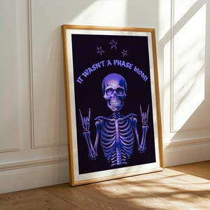 Pode incluir: Impressão de arte emoldurada com um esqueleto roxo fazendo um gesto de rock and roll. O texto "IT WASN'T A PHASE MOM!" está arqueado acima da cabeça do esqueleto, com três estrelas acima. A obra de arte está em um fundo preto.