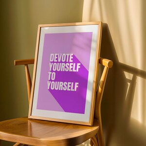 Può includere: Una stampa incorniciata con la scritta "DEVOTE YOURSELF TO YOURSELF" in lettere bianche su sfondo viola e lavanda. La cornice in legno è appoggiata su una sedia di legno, con ombre di luce solare.