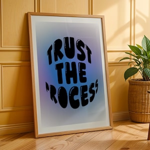 May include: A framed art print with the words "TRUST THE PROCESS" in black bubble letters against a blue and purple gradient background. The artwork is displayed on a wooden floor, leaning against a yellow wall. A potted plant in a woven basket is visible.