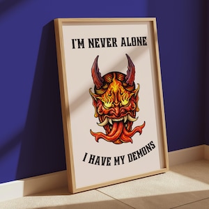 Puede incluir: Impresión artística enmarcada que presenta una ilustración detallada de una máscara de demonio con acentos de color naranja y amarillo fuego. La obra de arte incluye el texto "I'M NEVER ALONE I HAVE MY DEMONS" en negro, sobre un fondo crema.