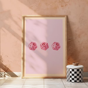 Puede incluir: Una impresión enmarcada con tres dados rosas, cada uno mostrando el número 6 y puntos en forma de corazón. Los dados están sobre un fondo rosa claro. El marco es dorado y la impresión está apoyada contra una pared color melocotón.