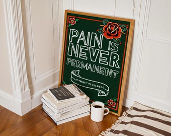 May include: A framed art print with the words "PAIN IS NEVER PERMANENT" in white block letters on a dark green background. The print features red roses and the phrase "BUT TONIGHT ITS KILLING ME". A stack of books and a coffee mug are in front of the print.