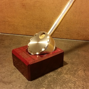 May include: A silver metal pipe with a round, ridged base and a long, straight stem. The pipe is sitting on a small, dark red wooden block.