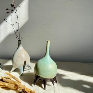 May include: Three ceramic vases with a unique, bulbous shape and three legs. The vases are in shades of green, blue, and brown.